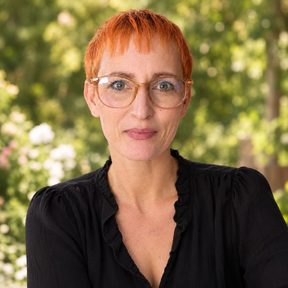 Porträtfoto einer Frau mit kurzen roten Haaren, Brille und schwarzer Bluse vor grünem Garten